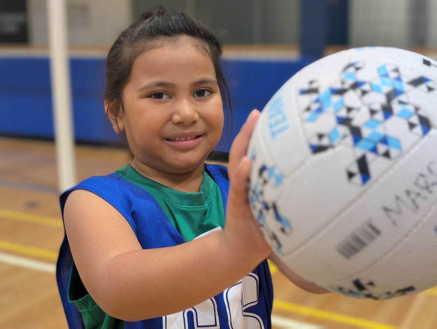 A smiling young girl wearing a blue sports bib holds a white, blue, and black netball forward while posing on an indoor timber court.